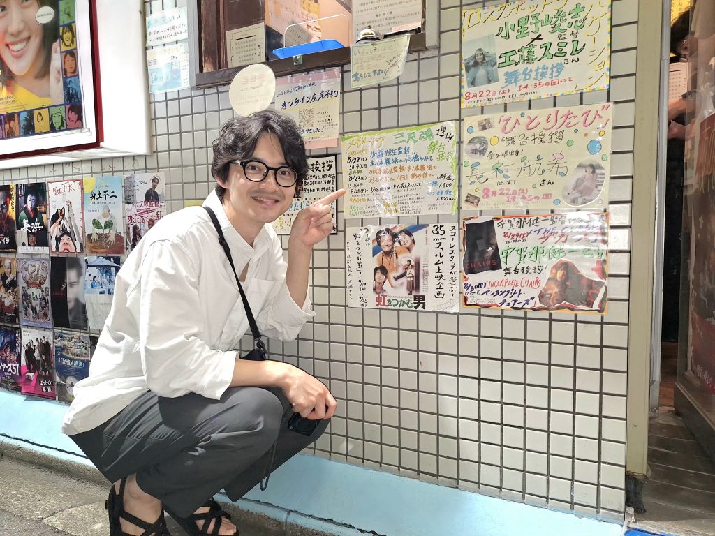 シネマスコーレの手書きの告知の横で撮影する木ノ本嶺浩さん